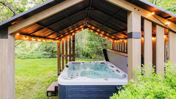 A hot tub under a wooden gazebo with string lights, surrounded by greenery. - Hot Tub / Home Theater / Private Acre / Walk to Beach / Family and Dog friendly (Bainbridge Island)