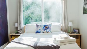 A bedroom with a large window, a bed with white linens, a blue floral pillow, and a bedside table with a lamp and a framed picture. - Hot Tub / Home Theater / Private Acre / Walk to Beach / Family and Dog friendly (Bainbridge Island)