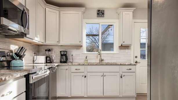 A modern kitchen with white cabinets, stainless steel appliances, and a window with a view of the outdoors. - Walk to Downtown [] Game Room [] Private Fenced Yard (Fruita)