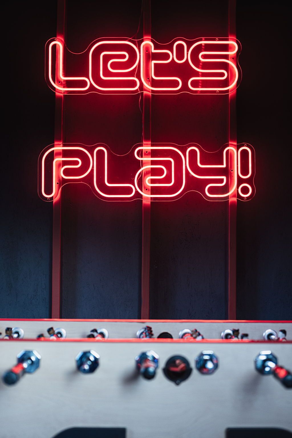 A neon sign that reads "LET'S PLAY!" with a foosball table below it.
