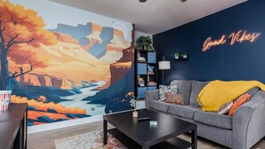 A living room with a mural of a canyon landscape, a gray sofa with a yellow throw, a black coffee table, and a neon sign that reads "Good Vibes". - Walk to Downtown [] Game Room [] Private Fenced Yard (Fruita)