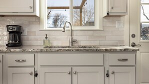 A kitchen with white cabinets, a granite countertop, a coffee maker, and a window with a view of trees. - Walk to Downtown [] Game Room [] Private Fenced Yard (Fruita)