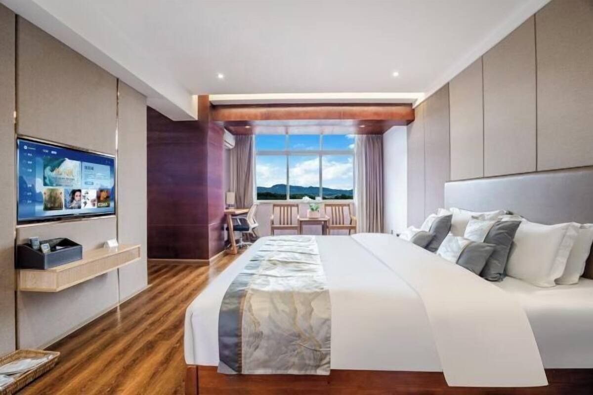 A hotel room with a large bed, a TV on the wall, a desk with a chair, and a window with a view.