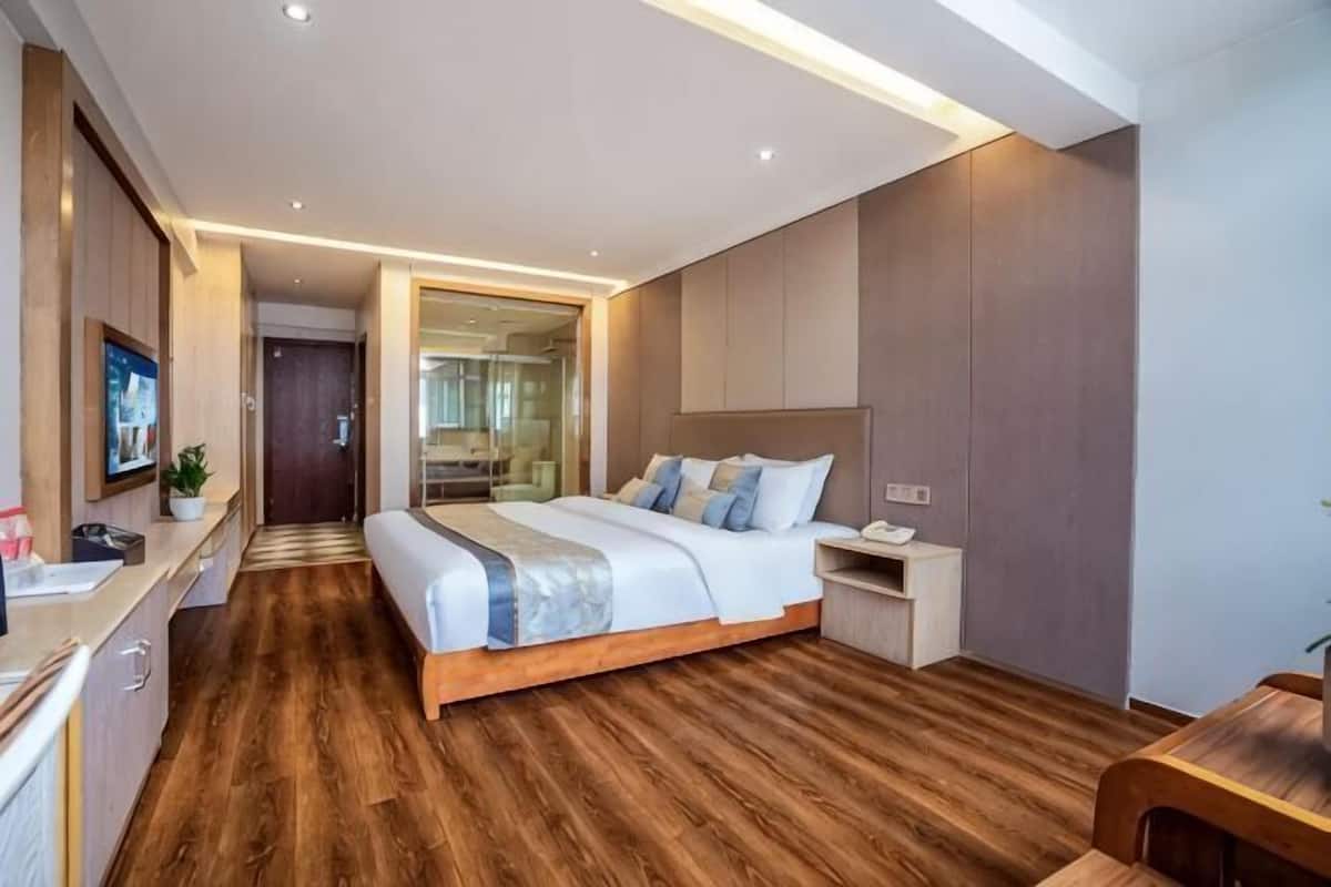 A modern hotel room with a wooden floor, a large bed with a headboard, a bedside table, a TV, and a bathroom visible through a glass door.