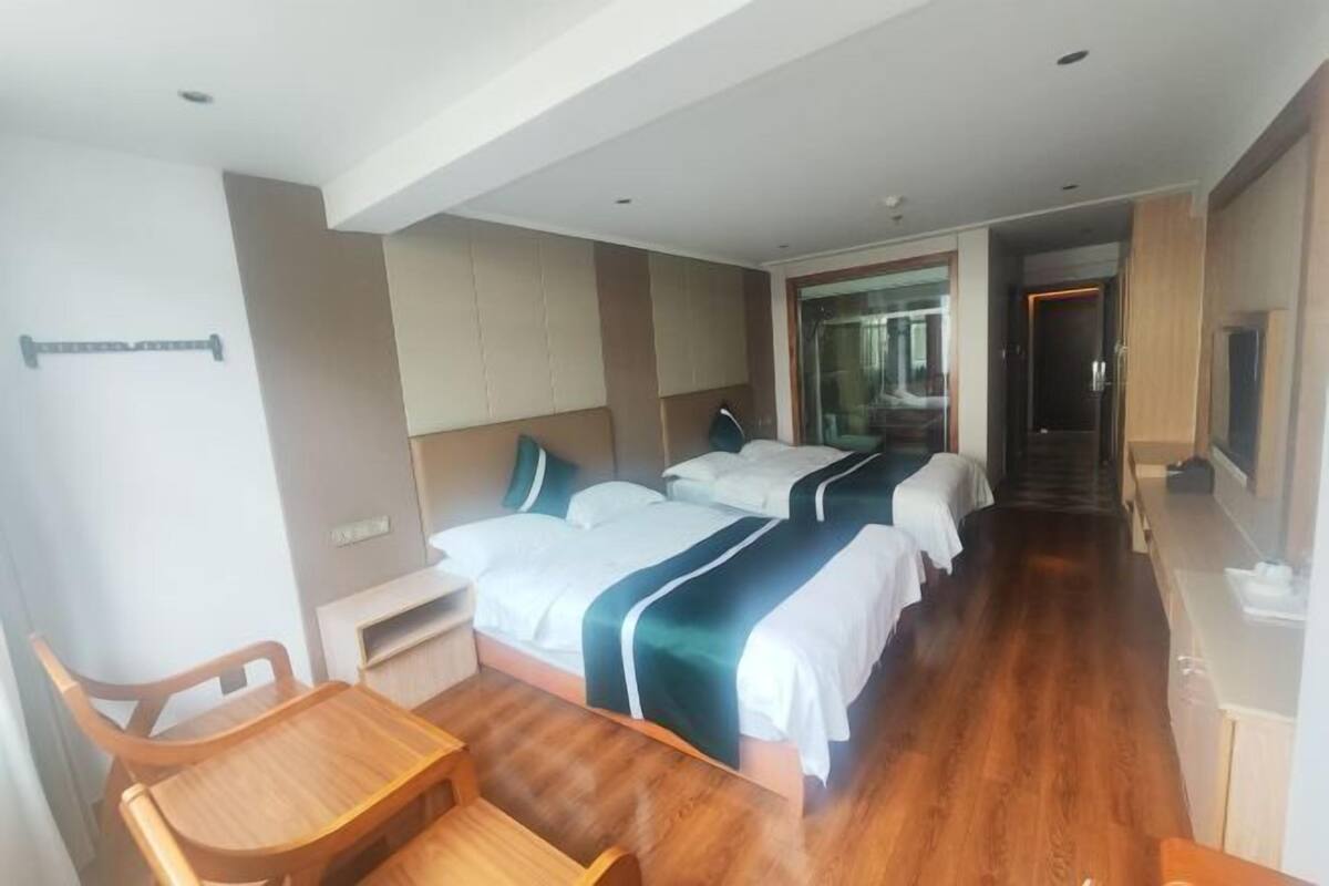 A hotel room with a wooden desk, two beds with white and green bedding, a large mirror, and a view of the hallway.