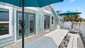 A wooden deck with white furniture and teal umbrellas, overlooking a teal building with large windows and doors. - 6 Bedroom + Crows Nest - Beach View (Galveston)
