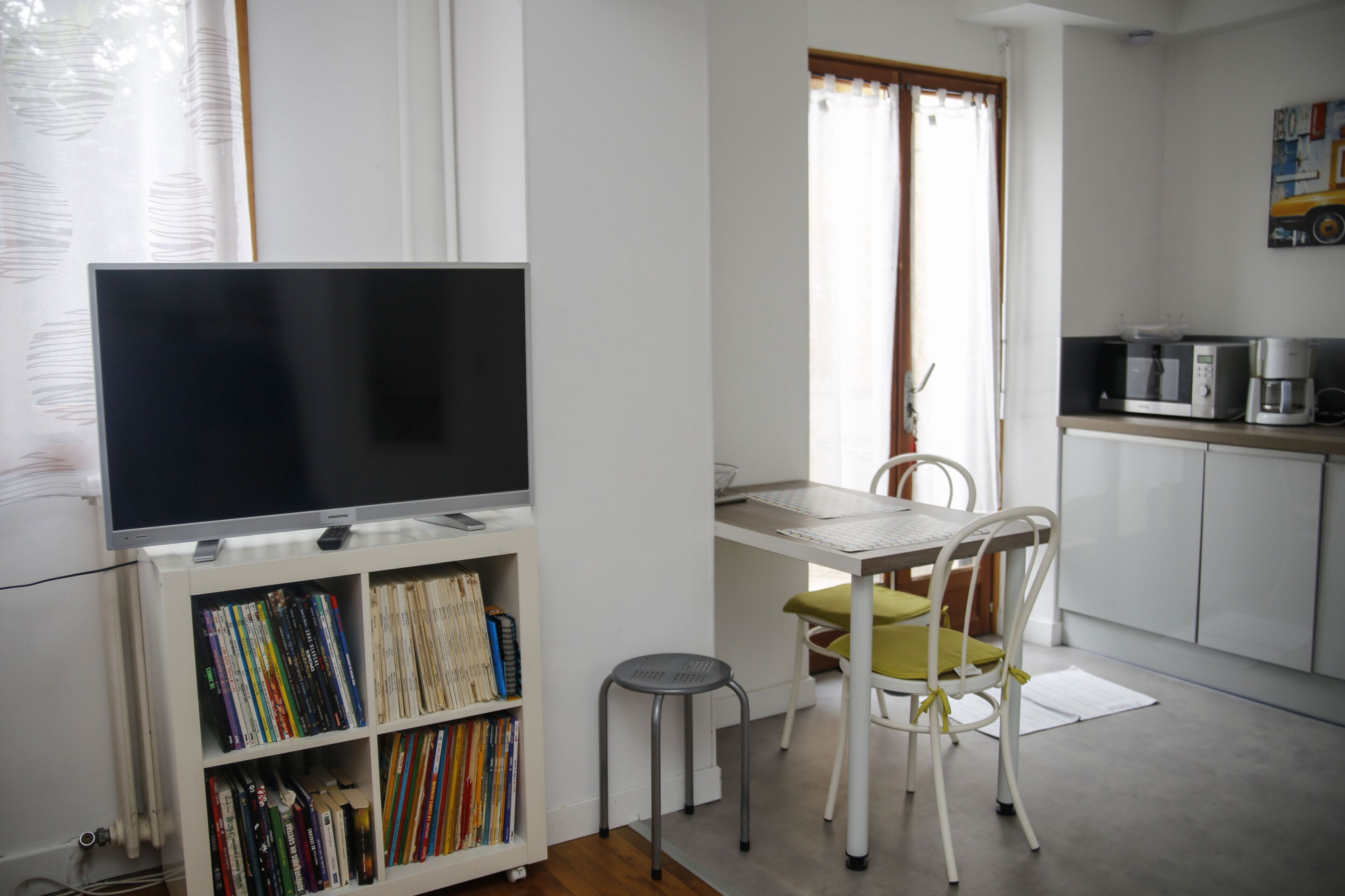 Un espacio habitable compacto con una televisión de pantalla plana sobre un mueble, una pequeña mesa de comedor con una silla, una kitchenette con microondas y una estantería llena de libros.