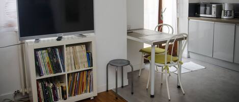 Un espacio habitable compacto con una televisión de pantalla plana sobre un mueble, una pequeña mesa de comedor con una silla, una kitchenette con microondas y una estantería llena de libros.