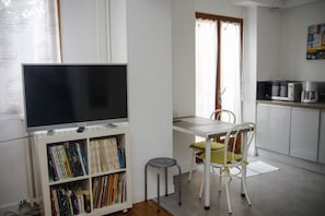A compact living space with a flat-screen TV on a stand, a small dining table with a chair, a kitchenette with a microwave, and a bookshelf filled with books.