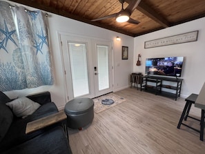 Un salon avec un canapé, un pouf et un téléviseur, le tout posé sur un plancher en bois.