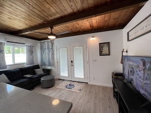A living room with a wooden ceiling, a black sofa, a coffee table, a TV, and a door with glass panels. - Lovely Private Kailua Cottage (Kailua)
