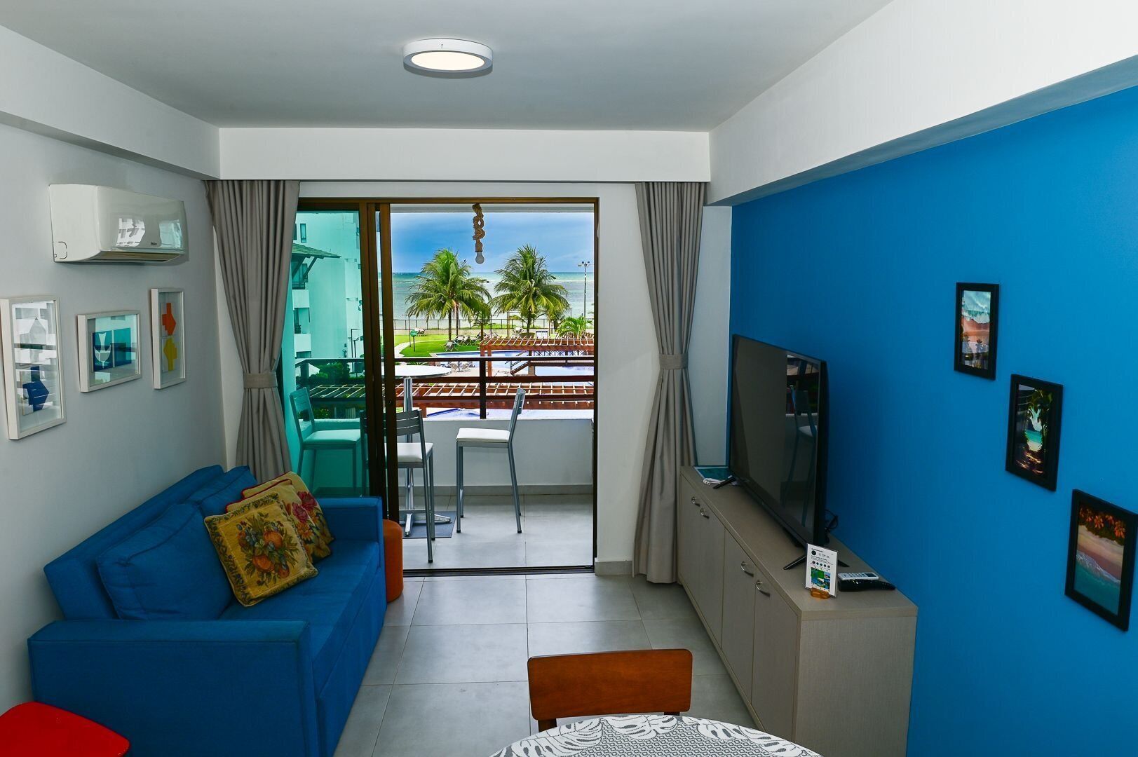 A modern living room with a blue sofa, a TV, and a balcony with a view of palm trees and the ocean.