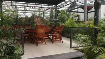 An outdoor patio with wicker furniture and a glass roof, surrounded by various plants.