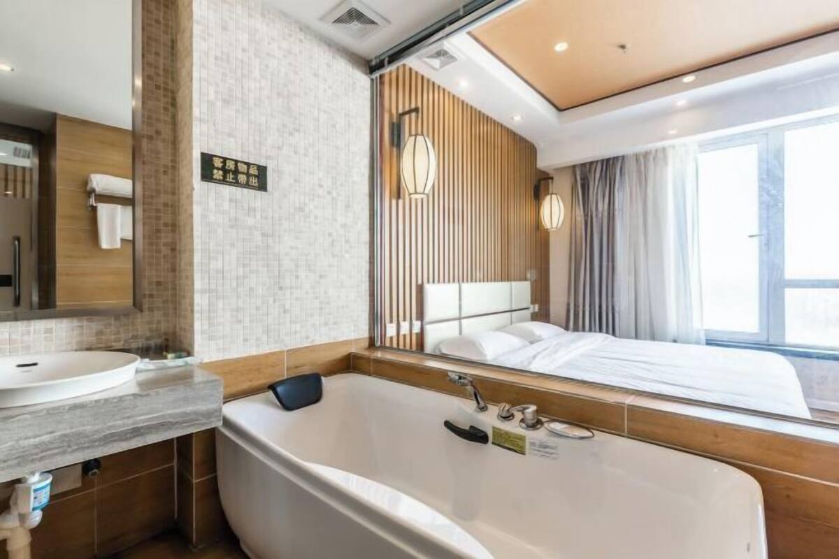 A modern hotel room with a large bathtub, a separate bathroom with a sink and mirror, a large bed, and a window providing a view.