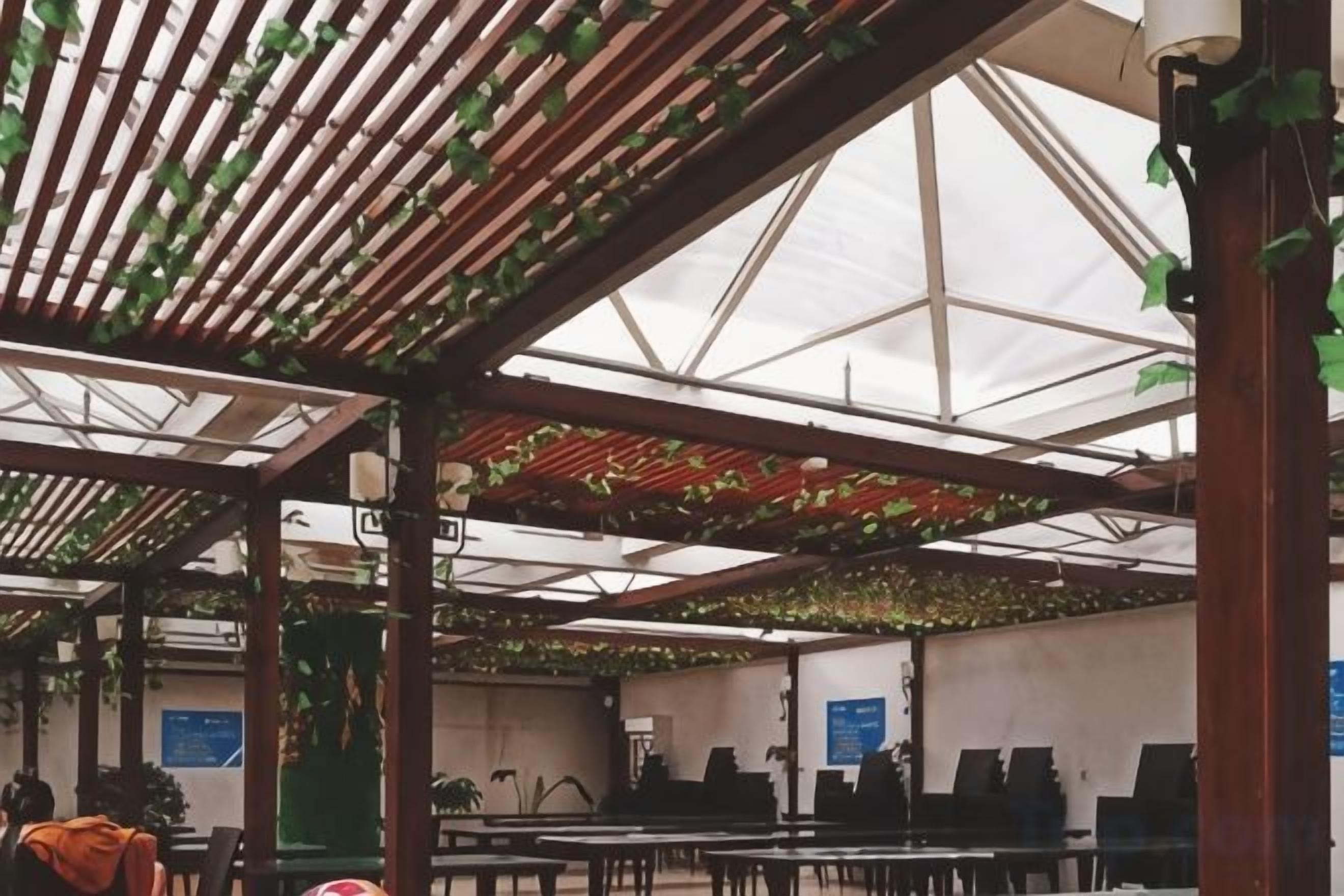 Un espacio interior con mesas y sillas, un techo alto con un diseño geométrico y plantas colgantes.