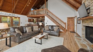 A spacious living area with leather sofas, a wooden coffee table, a stone fireplace, and a kitchen in the background. - Fireplace Deck, Game Room & Mountain Vibes (McHenry)