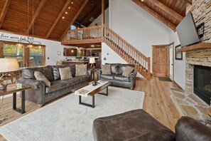A spacious living area with leather sofas, a wooden coffee table, a stone fireplace, and a kitchen in the background. - Fireplace Deck, Game Room & Mountain Vibes (McHenry)