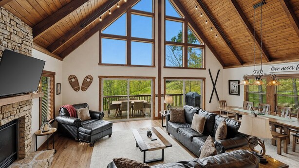 A spacious living area with a fireplace, leather sofas, a dining table, and large windows offering a view of the outdoors. - Fireplace Deck, Game Room & Mountain Vibes (McHenry)