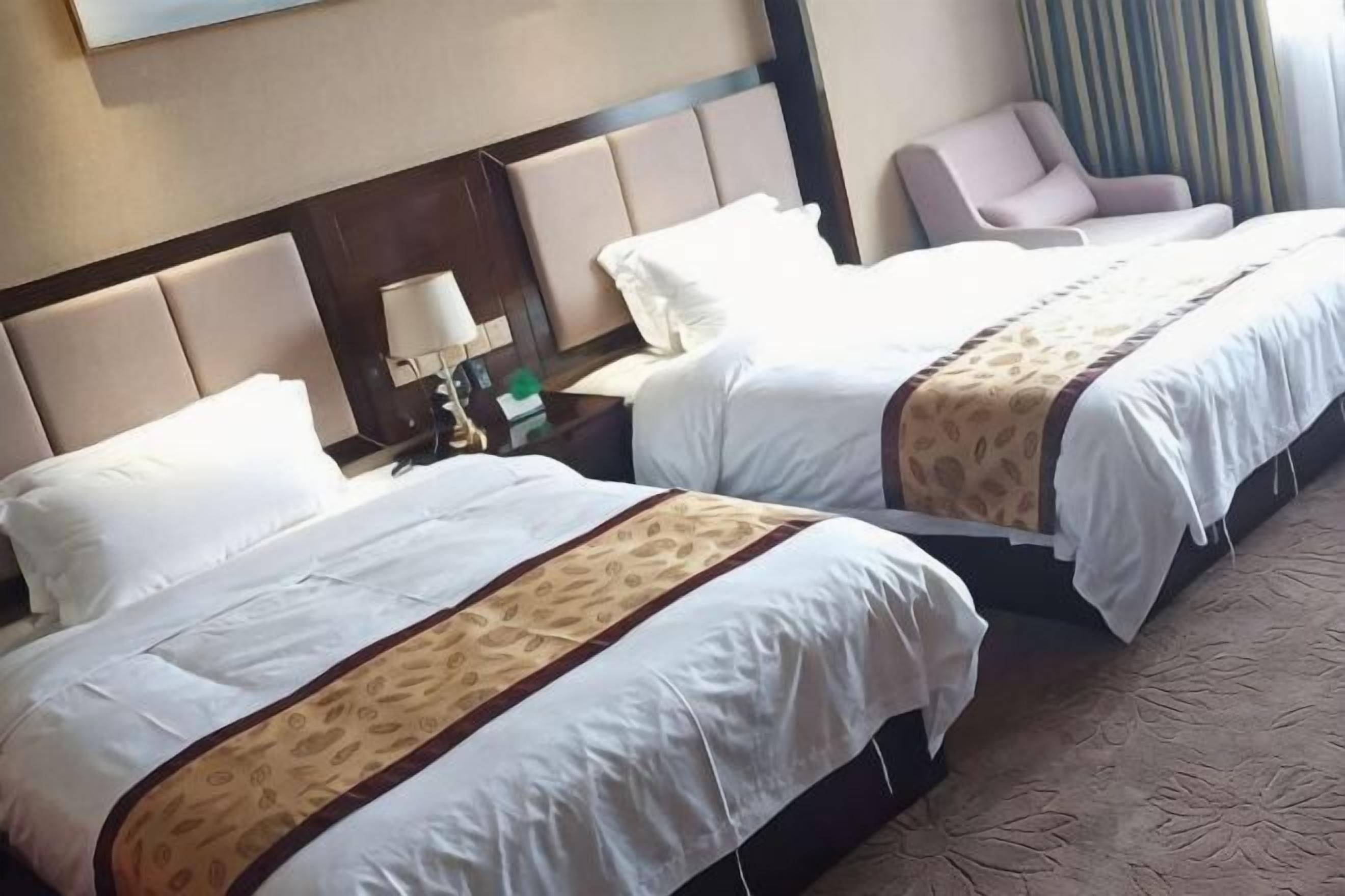Ein Hotelzimmer mit zwei Betten, einem Nachttisch mit Lampe, einem Sessel und Blick auf ein Gebäude.