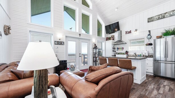 A spacious living area with a brown leather sofa, a white table with a lamp, and a kitchen with white cabinets and a stainless steel refrigerator. - Beautiful Beachfront Home with Amazing Views, Steps to Beach (Bay City)