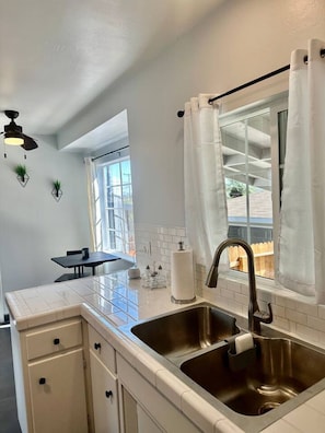 A modern kitchen with a stainless steel sink, white cabinetry, and a window with white curtains. - Bakersfield Sunset Stay (Bakersfield)