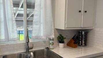 Une cuisine avec des armoires blanches, un évier en acier inoxydable, une fenêtre avec des rideaux blancs et une plante en pot posée sur le comptoir.