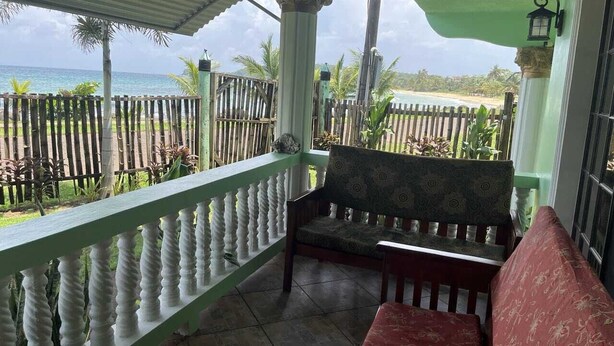 A covered outdoor seating area with a bench and a cushioned seat, overlooking a beach with a wooden fence and palm trees.