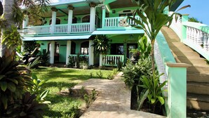 A two-story building with a balcony, surrounded by tropical plants and a concrete pathway.