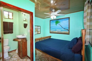A bedroom with a bed, a ceiling fan, a painting of a beach, and a bathroom with a toilet and sink.