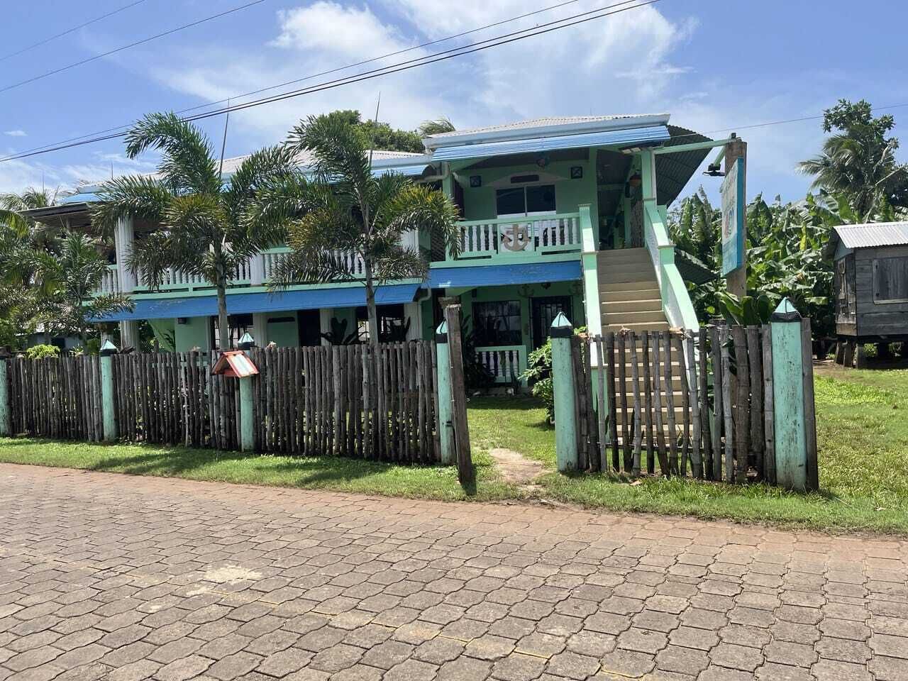 A two-story building with a blue roof and a green facade, surrounded by a wooden fence and palm trees.