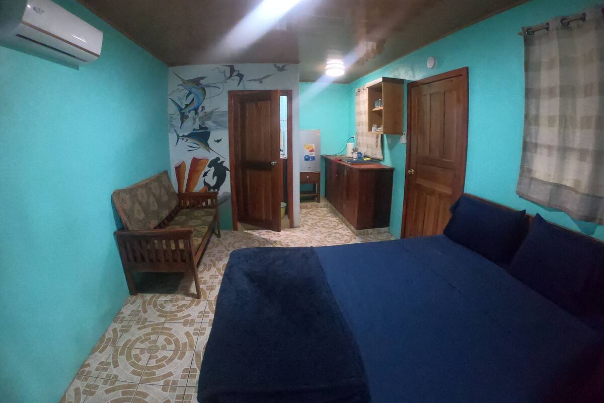 Une pièce aux murs de couleur turquoise, avec un lit, une chaise, une petite table et un mur orné d’une fresque.