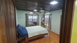 A bedroom with a bed, wooden flooring, a ceiling fan, and two windows with curtains.