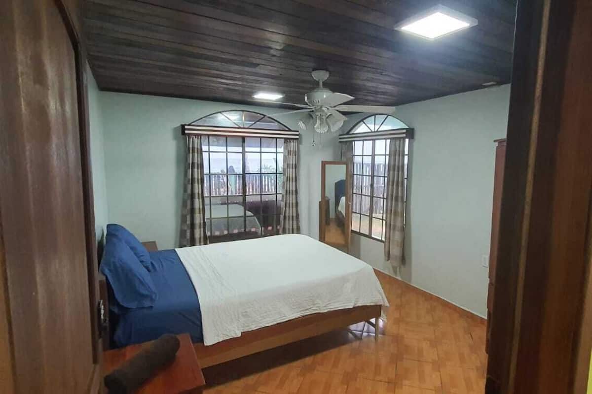 Une chambre à coucher avec un lit, un plancher en bois, un ventilateur de plafond et deux fenêtres avec des rideaux.