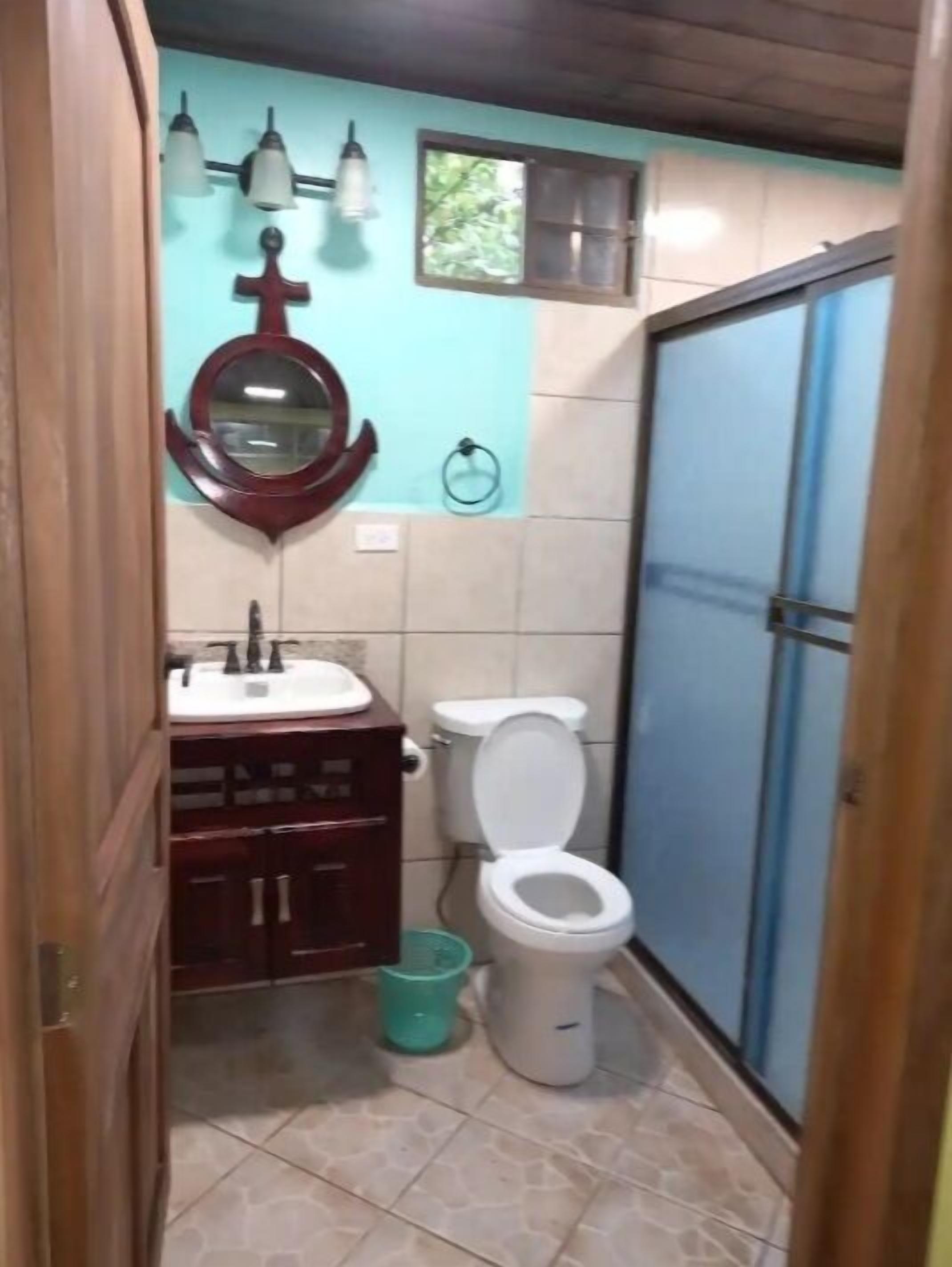 Un baño pequeño con inodoro, lavabo y espejo. Hay una ventana sobre el inodoro y una puerta a la derecha.