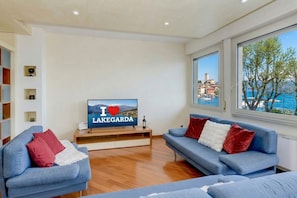 Un salon moderne avec un téléviseur affichant le message « J’♥ LAKEGARDA », une table basse en bois, deux canapés bleus ornés de coussins rouges et blancs, et une grande fenêtre offrant une vue sur une ville côtière et la mer.