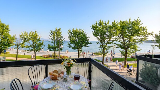 Une table dressée avec des assiettes, des verres et un vase de fleurs, sur un balcon donnant sur une plage où l’on voit des gens se promener et faire du vélo.