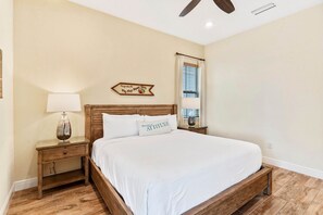 A bedroom with a wooden bed, a nightstand with a lamp, a window with blinds, and a wall sign.