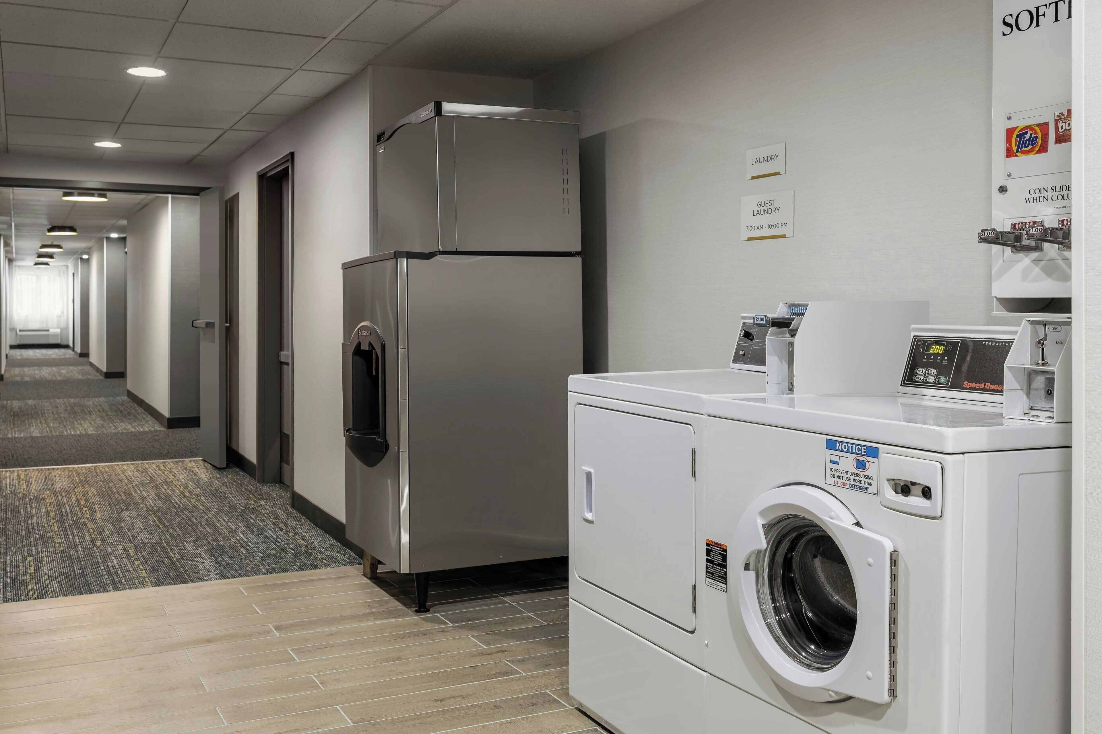 A hotel laundry room with a stainless steel refrigerator, a front-loading washing machine, and a coin-operated dryer.