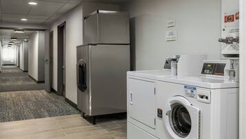 A hotel laundry room with a stainless steel refrigerator, a front-loading washing machine, and a coin-operated dryer.