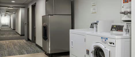 A hotel laundry room with a stainless steel refrigerator, a front-loading washing machine, and a coin-operated dryer.