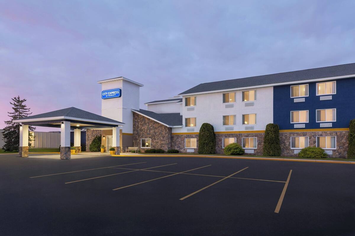 A hotel with a covered entrance, a parking lot, and a building with multiple windows and a stone facade.
