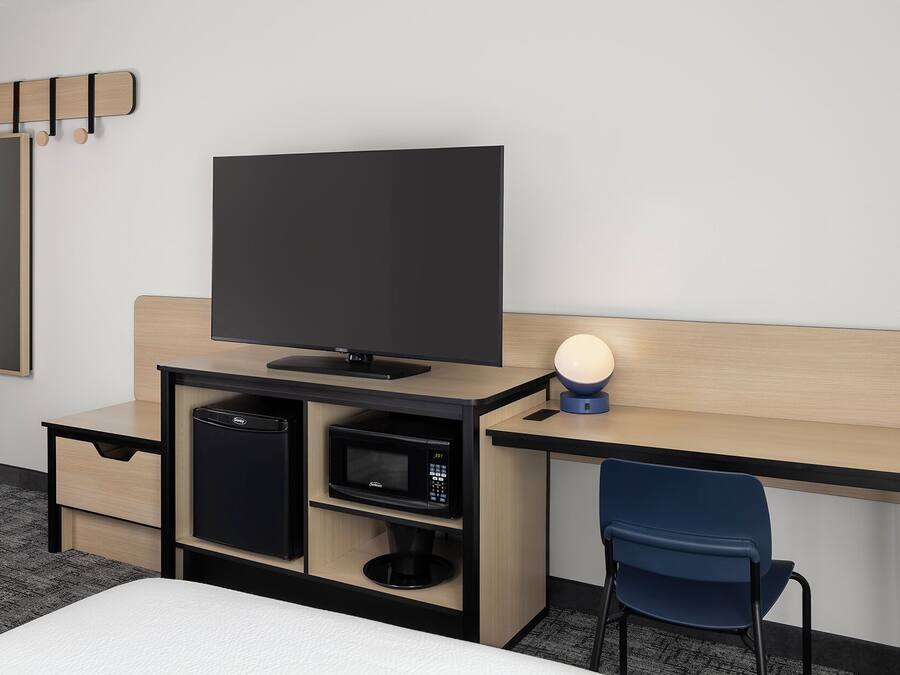 A hotel room with a bed, a desk with a flat-screen TV, a microwave, a small refrigerator, a chair, and a bedside lamp.
