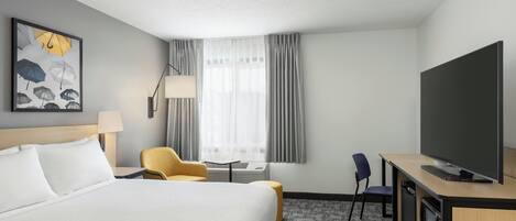 A hotel room with a bed, a desk with a flat-screen TV, a chair, a lamp, and a window with curtains.