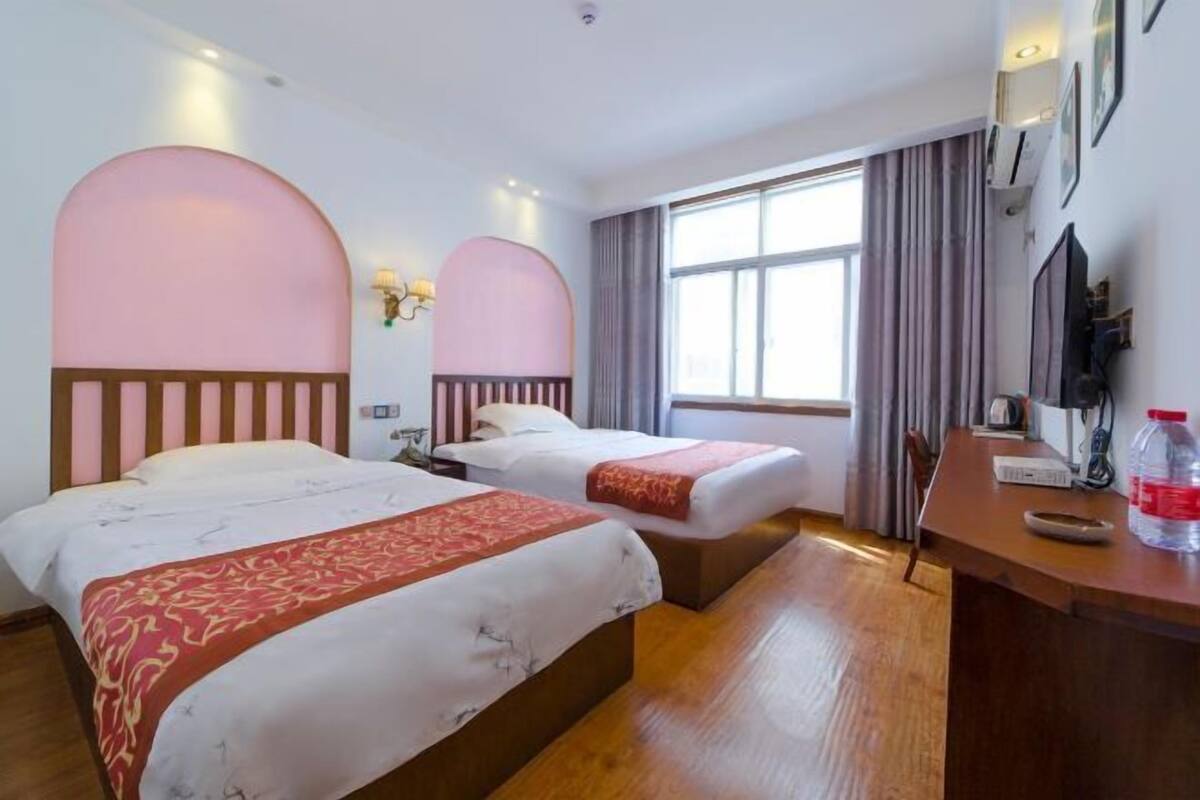 A hotel room with two beds, a desk, a TV, and a large window with curtains.