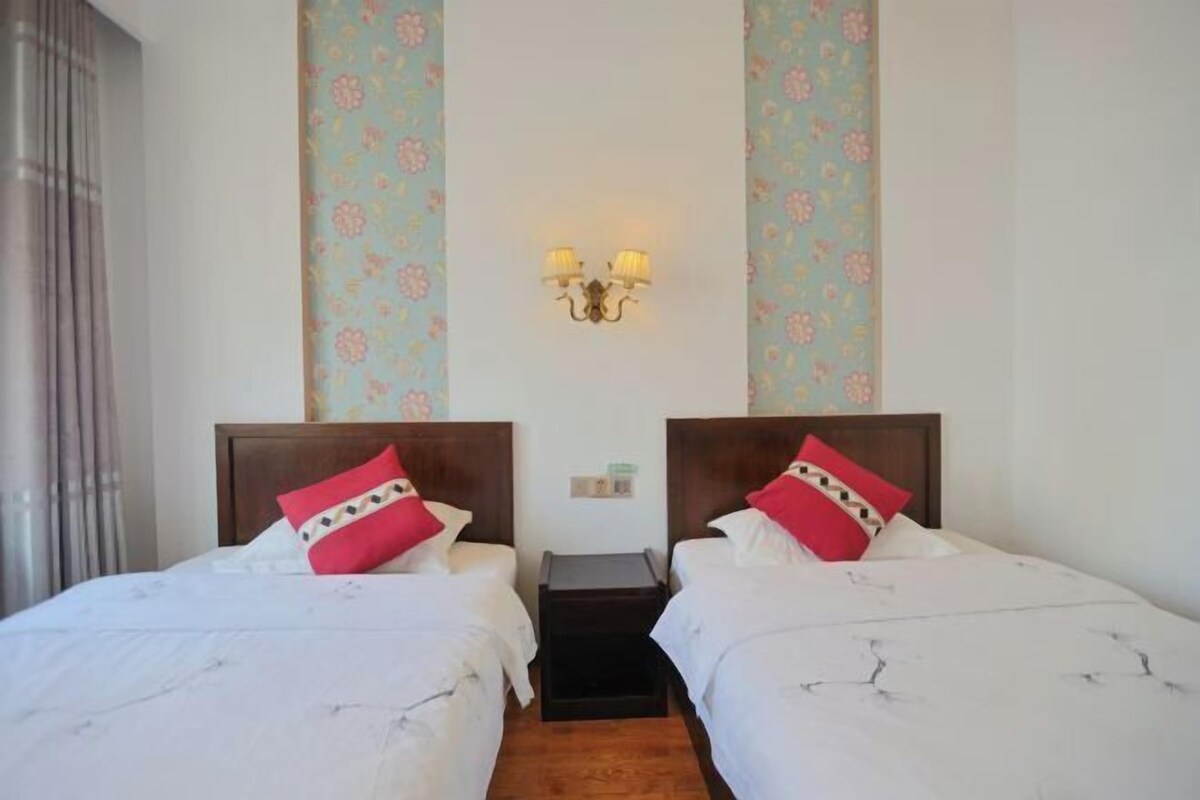 A hotel room with two beds, each with a wooden headboard, white bedding, and red decorative pillows. There is a small bedside table between the beds, and a wall-mounted light fixture above.