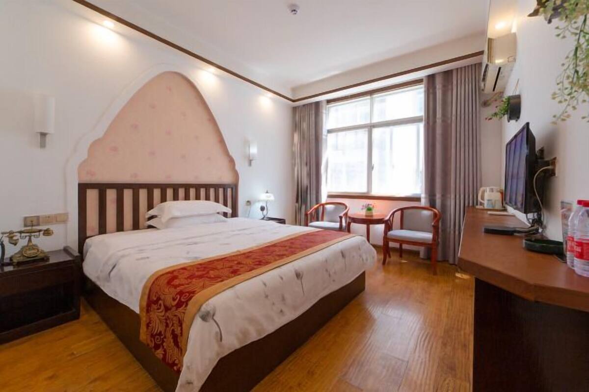 A hotel room with a large bed, a desk with a chair, a window with curtains, and a television on a stand.