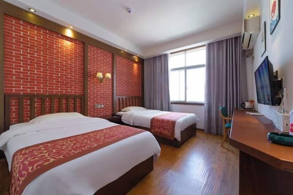 A hotel room with two beds, a wooden floor, a brick accent wall, a window with curtains, and a desk with a chair.