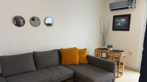 Een moderne woonkamer met een grijze hoekbank, twee mosterdgele kussens, een klein rond tafeltje met een plant en een wandairconditioner.