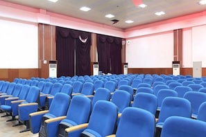 Ein Konferenzraum mit blauen Stuhlreihen, einem großen Bildschirm und Vorhängen.