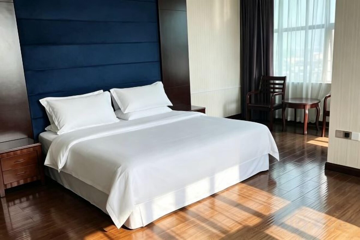 A neatly made bed with white linens in a room with wooden flooring and a dark blue headboard.